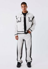 Chaqueta de mezclilla blanca con acentos de pintura en spray negra, combinada con jeans blancos a juego que tienen rayas laterales negras. Camiseta negra debajo.