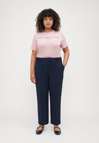 Hellrosa Baumwoll-T-Shirt mit marineblauem Text, kombiniert mit hochtaillierten, weit geschnittenen marineblauen Hosen. Das Model steht mit den Händen in den Taschen.