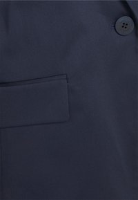 Tumma laivastonsininen takkimateriaali, jossa on läpällinen tasku vasemmalla puolella ja suuri musta ommeltu nappi kauluksen lähellä.