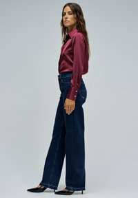 Camisa de satén burdeos con mangas largas y detalles de botones; combinada con jeans de pierna ancha de color azul oscuro y tacones negros de punta.