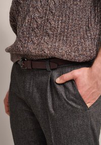 Brown and navy knitted sweater with a textured pattern, paired with gray textured trousers. A dark belt is worn, hand rests in pocket.