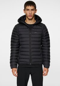 Svart vadderad jacka med dragkedja framtill och huva. Har en kviltad design och en diskret logotyp på bröstet. Texturerat, lättviktigt material.