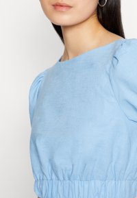 Top de linho azul claro com mangas curtas bufantes, decote redondo e detalhe na cintura franzido. Textura suave com um acabamento delicado.