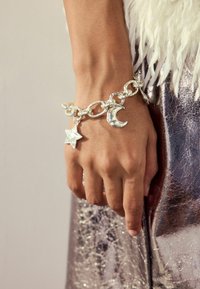 Sølv charmsarmbånd med sammenkoblede kæder, der har stjerne- og halvmånedecharm. Hånd hvilende mod en skinnende metallisk overflade.