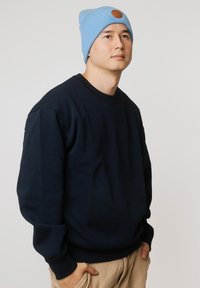 Granatowa bluza z prążkowaniem na mankietach i u dołu. Jasnoniebieska czapka beanie z brązową skórzaną naszywką. Dopasowanie casual. Gładkie tło.