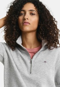Grijze zip-up sweatshirt met hoge kraag, voorzien van een klein geborduurd logo op de borst en gedragen over een roze t-shirt. Gestructureerde stof.