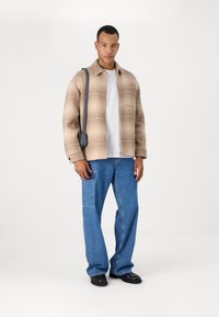 Jachetă plaid bej, tricou alb, blugi ușor evazați albastre și încălțăminte neagră. Jacheta are guler și buzunare frontale.