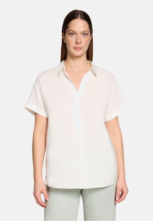 Frau mit glatten braunen Haaren trägt eine weiße, kurzärmelige, strukturierte Bluse und hellgraue Hose, stehend vor einfarbig hellem Hintergrund.