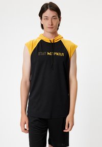 Musta ilmanhiha huppari keltaisilla yksityiskohdilla ja naruilla. Edessä teksti "STAY MOTIVATED". Sileä kangas, urheilullinen istuvuus.
