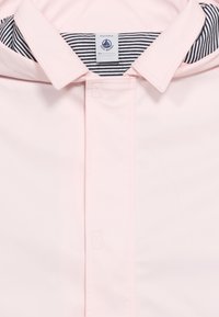 Col col et rabat avant d'un imperméable rose clair avec doublure intérieure rayée noire et blanche et étiquette de taille Petit Bateau.