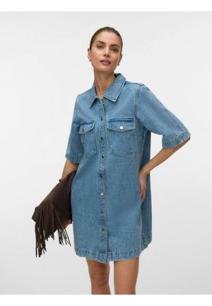 Nő rövid ujjú kék farmer gombos ruhában, barnás rojtos borítéktáskát tartva, egyik kezével a csípőjén.