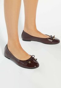 Brown patent leather ballet flats with a rounded toe, featuring a bow accent and decorative beads. Flat sole and smooth texture.