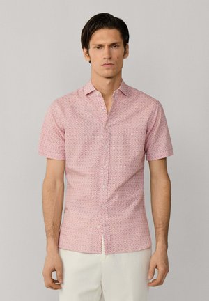 Hombre con camisa rosa de manga corta y botones, con patrón de diamantes y pantalones blancos, de pie frente a un fondo claro y liso.