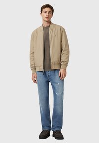 Chaqueta bomber beige sobre un suéter verde oscuro, combinada con jeans claros desgastados y zapatos gruesos marrón oscuro.