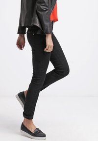Vaqueros skinny negros con una textura suave, de cintura alta y cremallera lateral. Combinados con una chaqueta de cuero y zapatos cómodos tipo slip-on.