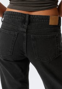 Suurendatud pilt alaseljast ja puusadest, seljas kõrge vöökohtadega mustad denimid, millel on tagataskud ja pruun nahast brändipatch.