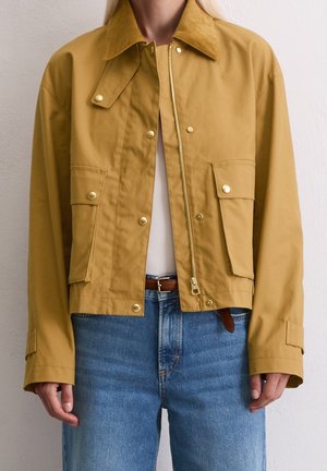 Mujer con una chaqueta amarilla mostaza con botones a presión y cremallera, dos bolsillos frontales con solapa, combinada con vaqueros azules y top blanco.