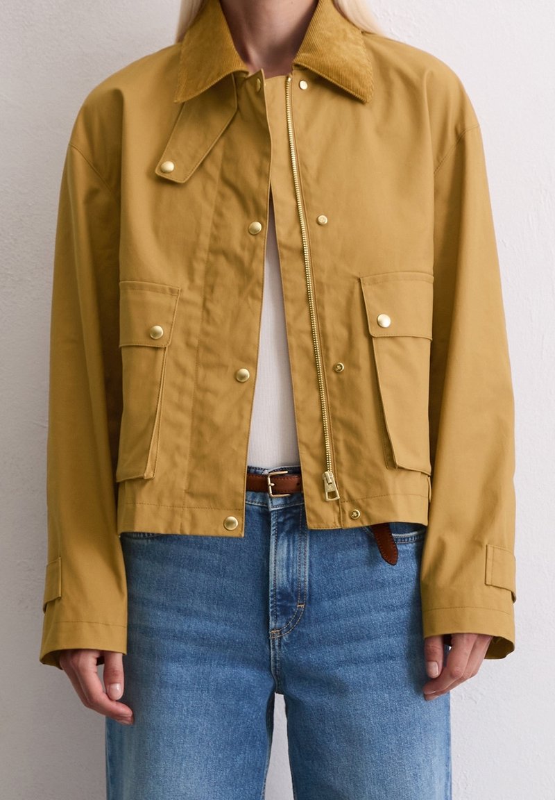 Femme portant une veste jaune moutarde avec boutons-pression et fermeture éclair, deux poches avant à rabat, associée à un jean bleu et un haut blanc.