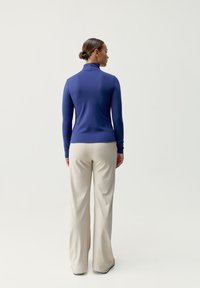 Långärmad topp med hög hals i enfärgad blå tyg, kombinerad med vida ljusbeiga byxor. Outfiten har en figursydd silhuett.