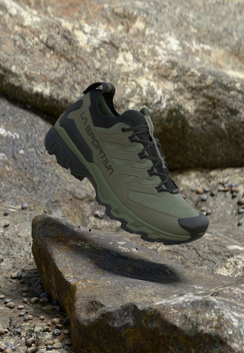Olivgrüner und schwarzer La Sportiva Wanderschuh in der Luft über felsigem Gelände, zeigt robuste Sohle und seitliches Logo.