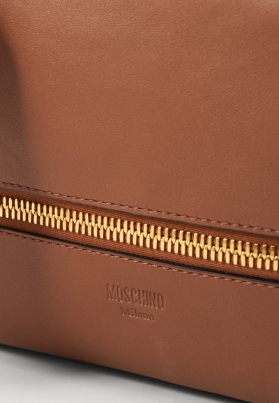 Barna bőrtáska, melynek kiemelkedő arany cipzára és a "Moschino Milano" felirattal ellátott domború logója a táska alsó részén található. Sima textúra, strukturált dizájn.