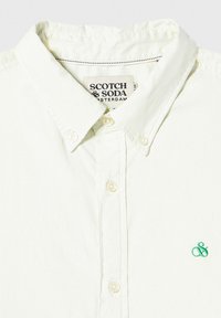Lys Gul skjorte med knapper og krave, der har et grønt broderet logo på brystet og kreme farvede knapper.