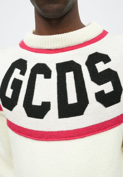 Baltas megztinis su dideliu juodu "GCDS" logotipu, raudonu akcentu aplink iškirpę ir raištiniu raštu.