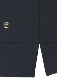 Navyblauw top met een ronde, gouden embleem aan de linkerkant, een soepele textuur en een gespleten zoomdetail onderaan voor een gelaagde uitstraling.