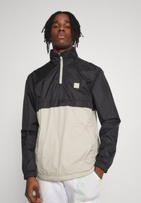 Giacca leggera a pullover con parte superiore nera e parte inferiore beige, dotata di mezza cerniera, polsini elasticizzati e un piccolo patch con logo.