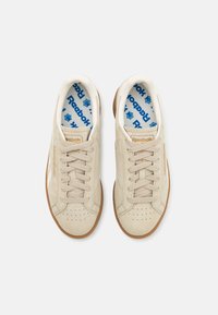 Beige mockaskor med en texturerad yta, rund tå och tanfärgad gummisula. Blå tryckta innersulor med "Reebok"-logotyp.