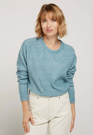 Pull bleu clair avec col rond, poignets côtelés et texture douce. Présente des détails de couture subtils le long des épaules. Associé à un pantalon crème.