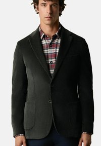 Ciemnozielona marynarka z kordura o dopasowanym kroju, z dwoma przednimi kieszeniami i pojedynczym zapięciem na guzik, noszona na koszuli w kratę.