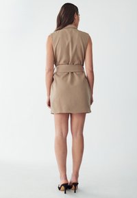 Beiges, ärmelloses Kleid mit Gürtel, das einen Kragen und eine glatte Textur aufweist. Der Saum ist über dem Knie, von hinten gezeigt.