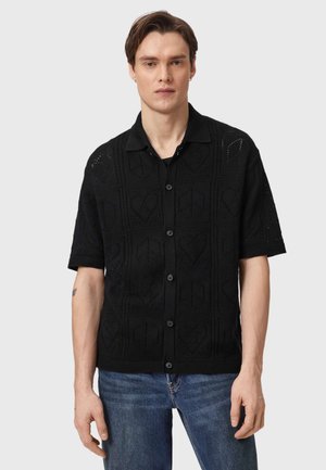 Young man wearing a black short-sleeve button-up shirt with heart and peace symbol patterns, paired with blue jeans, standing against gray background.