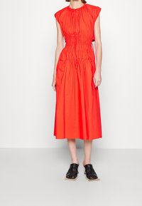 Kvinna i en ärmlös, ljusröd midiklänning med draperade detaljer och svarta klacksandaler, stående mot en vit vägg.