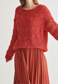 Czerwony sweter dzianinowy z teksturowanym wzorem w zygzak, posiadający szeroką dekolt i długie rękawy, w zestawieniu z plisowaną spódnicą w kolorze rdzy.