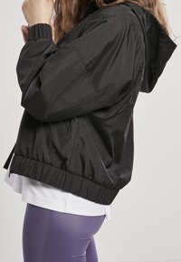 Czarna kurtka przeciwwiatrowa z nylonu z kapturem, elastycznymi mankietami i krótkim krojem, w połączeniu z białą, warstwową bluzką i fioletowymi legginsami.