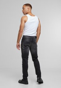 Mann i hvit tank topp og svarte jeans som står og ser over venstre skulder foran en enkel grå bakgrunn.