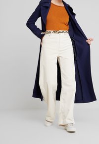 Marineblauer Trenchcoat über einem orangefarbenen Oberteil, kombiniert mit weißen Cordsamt-Hosen mit weitem Bein und einem beigen Gürtel im Leopardenmuster. Weiße Schuhe.
