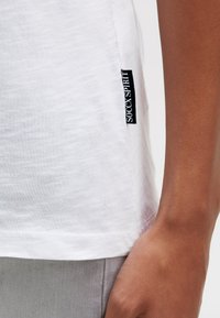 Weißes T-Shirt aus strukturiertem Stoff, mit einem Seitenschild mit der Aufschrift "SOCX SPIRIT." Der Saum ist mit einer genähten Kante versehen. Graue Hose teilweise sichtbar.