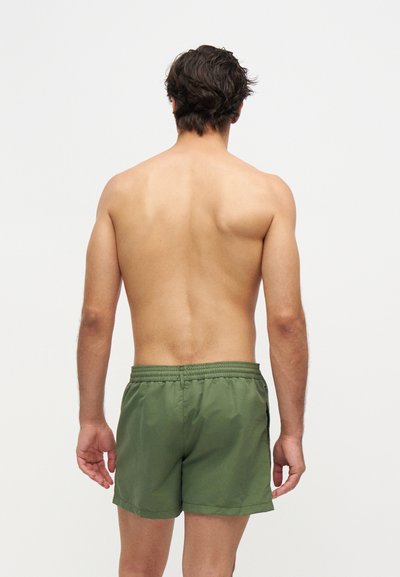 Homme aux cheveux foncés, debout torse nu, de dos, portant un short vert sur un fond blanc uni.