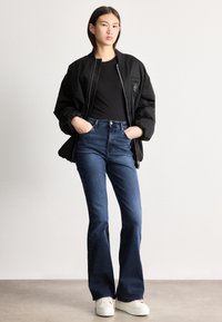 Sieviete, kas valkā melnu bombera stila jaku pār melnu topu, zilas augsta vidukļa zvana formas džinsas un baltus sporta apavus, stāvot ar rokām kabatās.