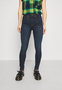Jeans skinny de color azul oscuro con una textura suave, diseño de cinco bolsillos y costuras en contraste, combinados con zapatos negros que presentan adornos de flecos.