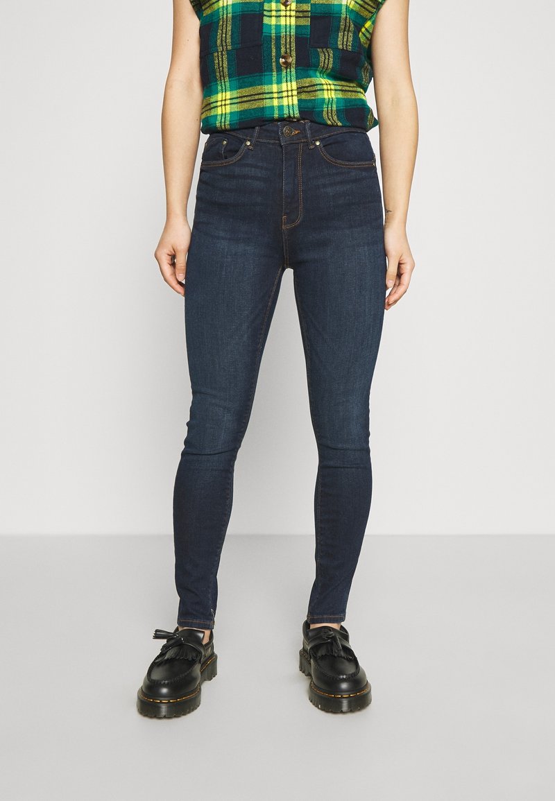 Jeans skinny de color azul oscuro con una textura suave, diseño de cinco bolsillos y costuras en contraste, combinados con zapatos negros que presentan adornos de flecos.