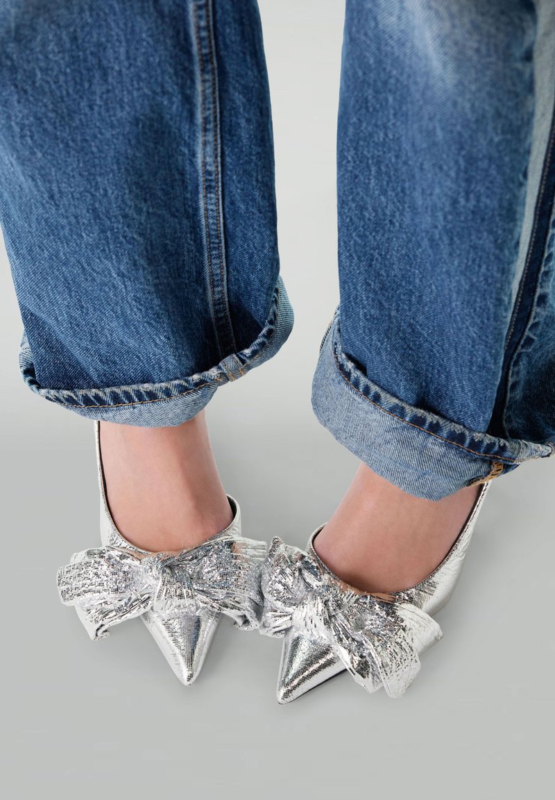 Chaussures à talons hauts argentées avec des pointes fines, agrémentées d'un nœud texturé sur le devant, associées à un jeans en denim retroussé.