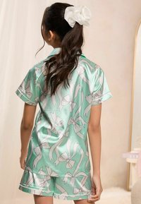 Conjunto de pijama de satén verde menta con mangas cortas y pantalones cortos, que presenta un patrón de cintas y lazos blancos. Textura suave, corte relajado.