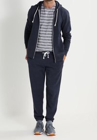 Sudadera navy con cordones blancos sobre una camiseta de rayas gris y blanca, combinada con joggers navy a juego y zapatillas deportivas grises.