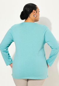 Maglia a maniche lunghe turchese in morbido tessuto. Caratterizzata da un colletto rotondo e un taglio dritto con un orlo posteriore più lungo.