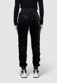 Pantaloni della tuta neri in velluto con una finitura lucida e testurizzata, caratterizzati da un audace motivo logo a copertura totale e polsini a coste; abbinati a sneaker bianche.