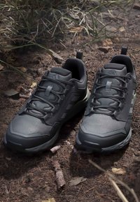 Paire de chaussures de randonnée athlétiques gris foncé avec lacets reposant sur un sol en terre avec des feuilles sèches et de l'herbe éparpillées.